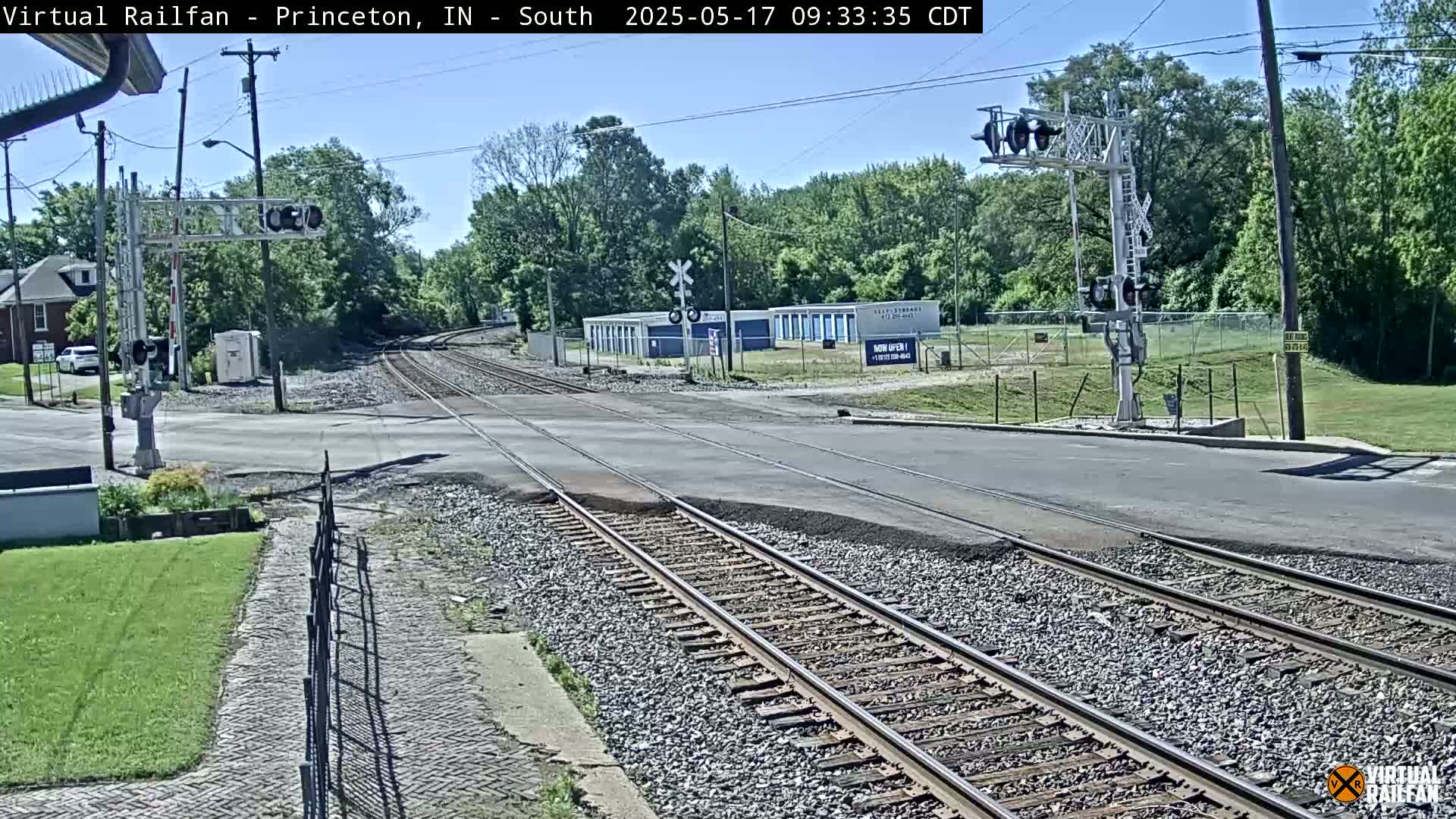 A sunny day shows a railroad crossing with train tracks intersecting a road, surrounded by greenery and a storage facility.