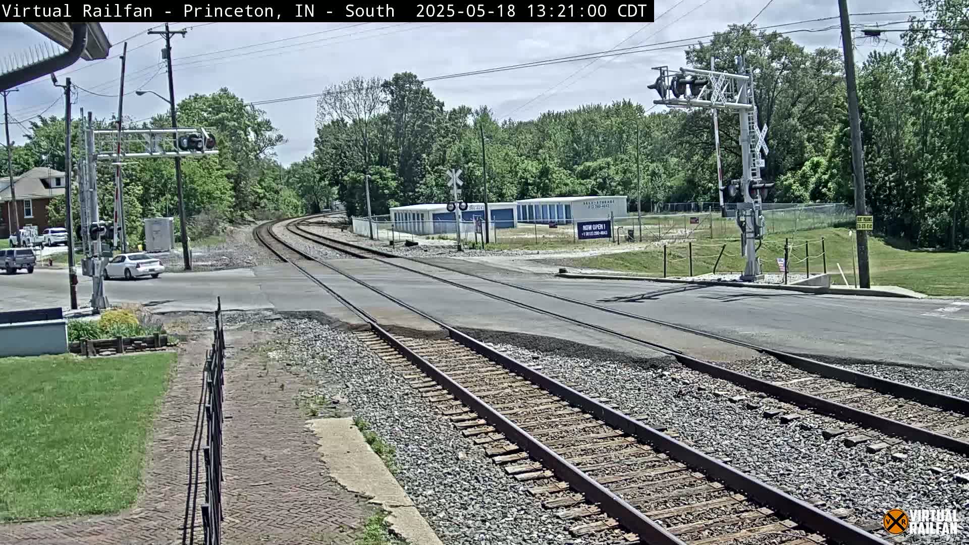 A sunny day shows a railroad crossing with two sets of train tracks intersecting a road, several cars are visible near the crossing, and there are trees and buildings in the background.