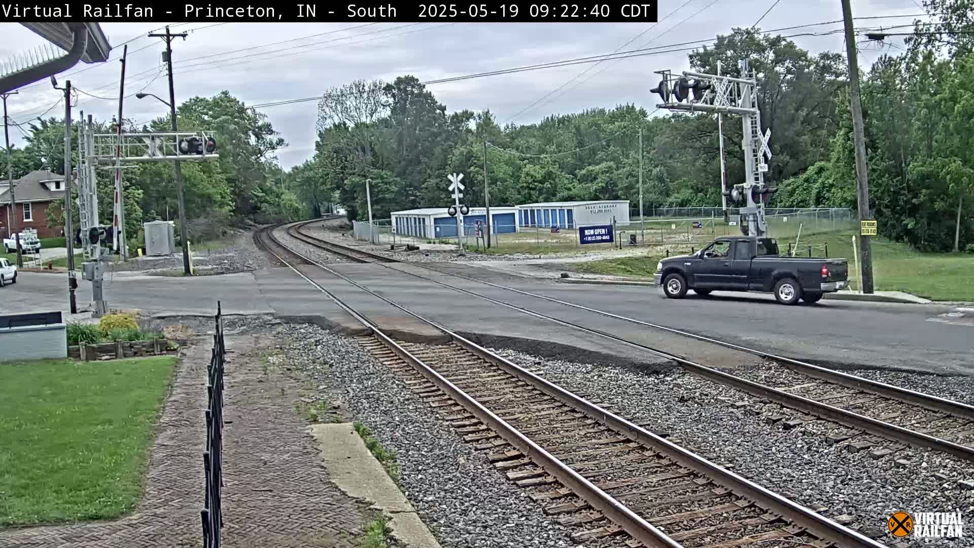A dark-colored pickup truck drives across a railroad crossing on a partly cloudy day, with train tracks extending into a tree-lined area.