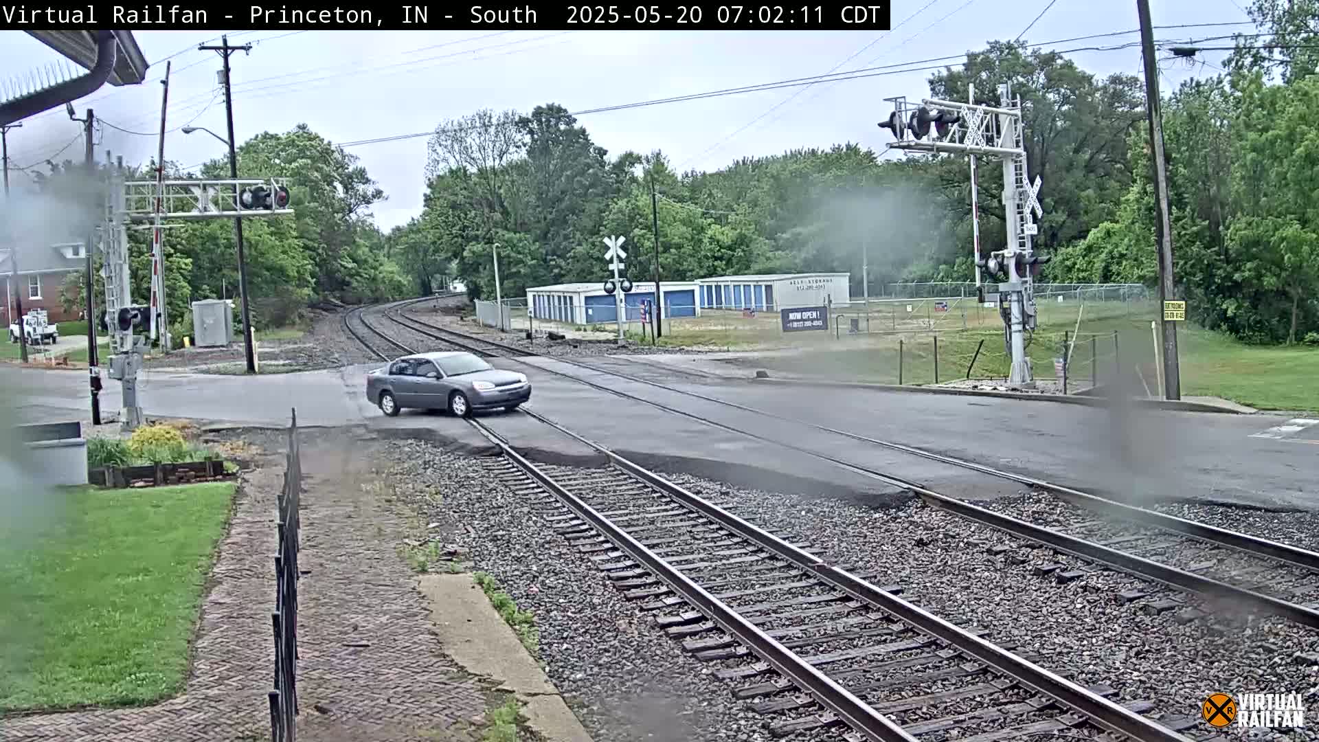 A gray car crosses a railroad track at a road intersection on an overcast day.