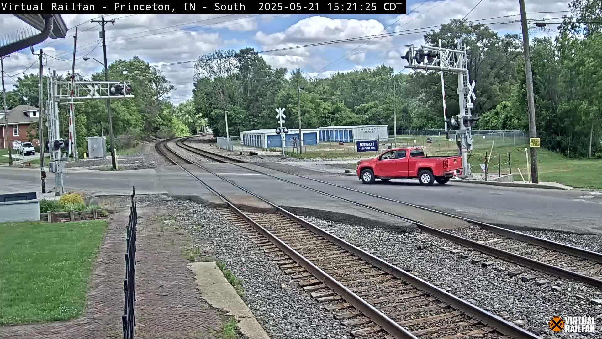A red pickup truck is crossing a road with railroad tracks under partly cloudy skies, near a row of storage units and some trees.