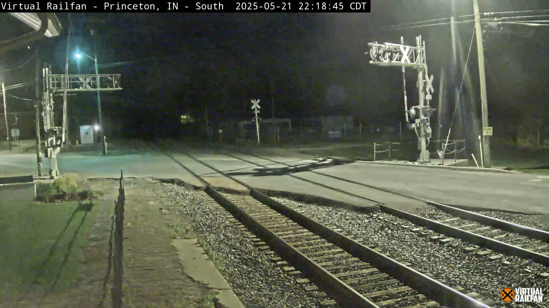 A nighttime view of a railroad crossing with two sets of tracks intersecting a road under artificial lighting.