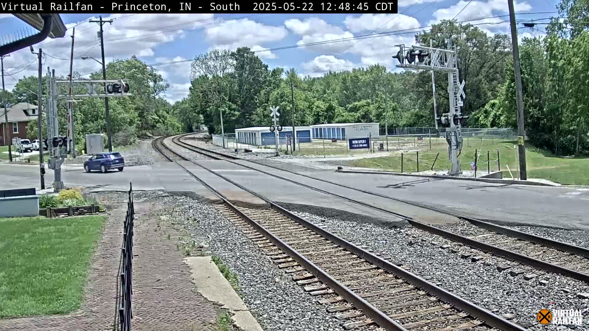 A partly cloudy day shows a railroad crossing with a single dark-colored SUV stopped at the intersection, surrounded by trees and a storage facility.