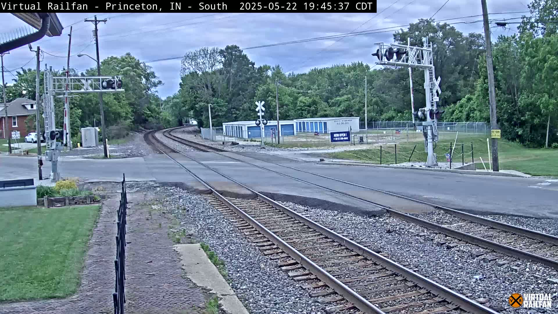 A railroad crossing with train tracks intersecting a road under an overcast sky, surrounded by trees and some buildings.