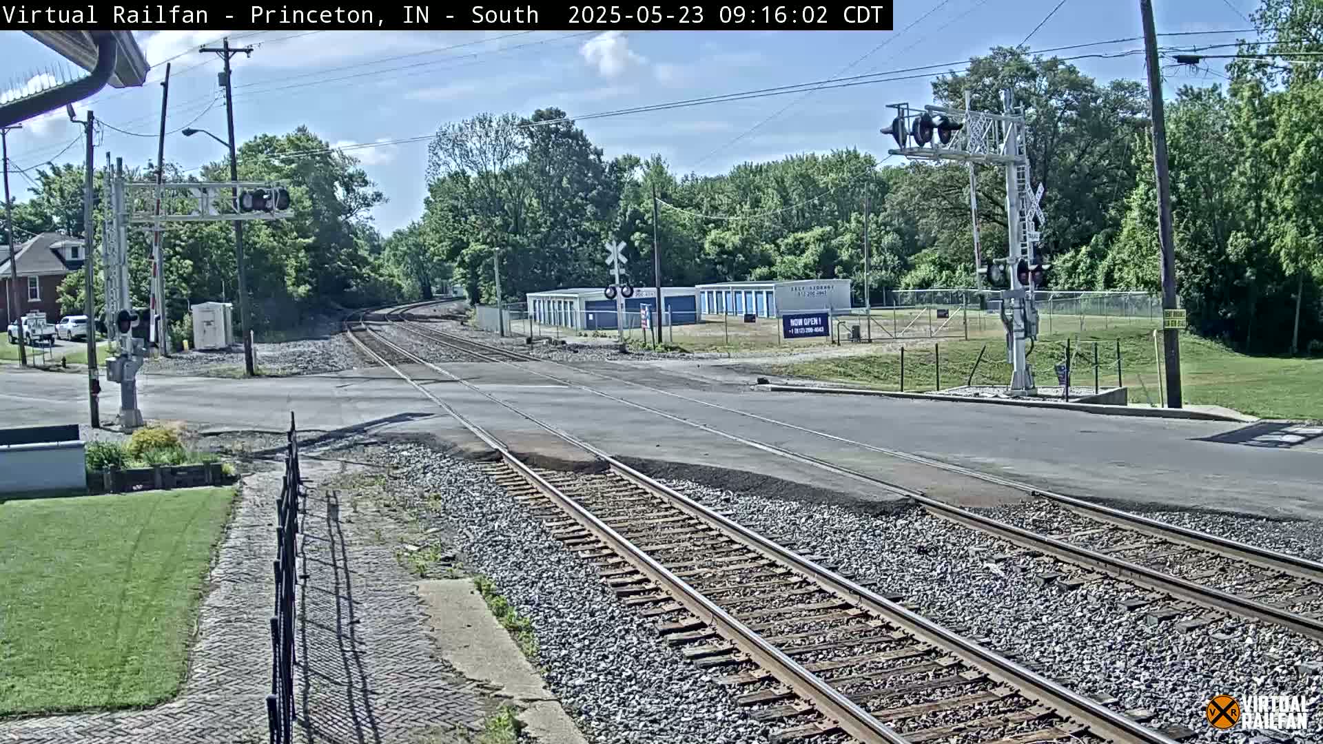 A railroad crossing with train tracks intersecting a road, under a mostly sunny sky, is surrounded by trees and buildings.