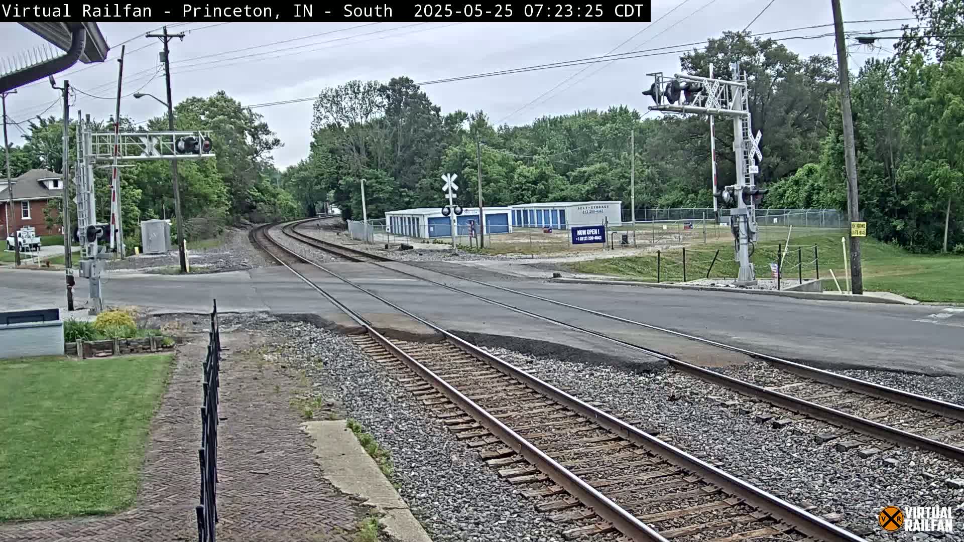 A railroad crossing with two sets of tracks intersects a road under an overcast sky, bordered by trees, grass, and storage units.