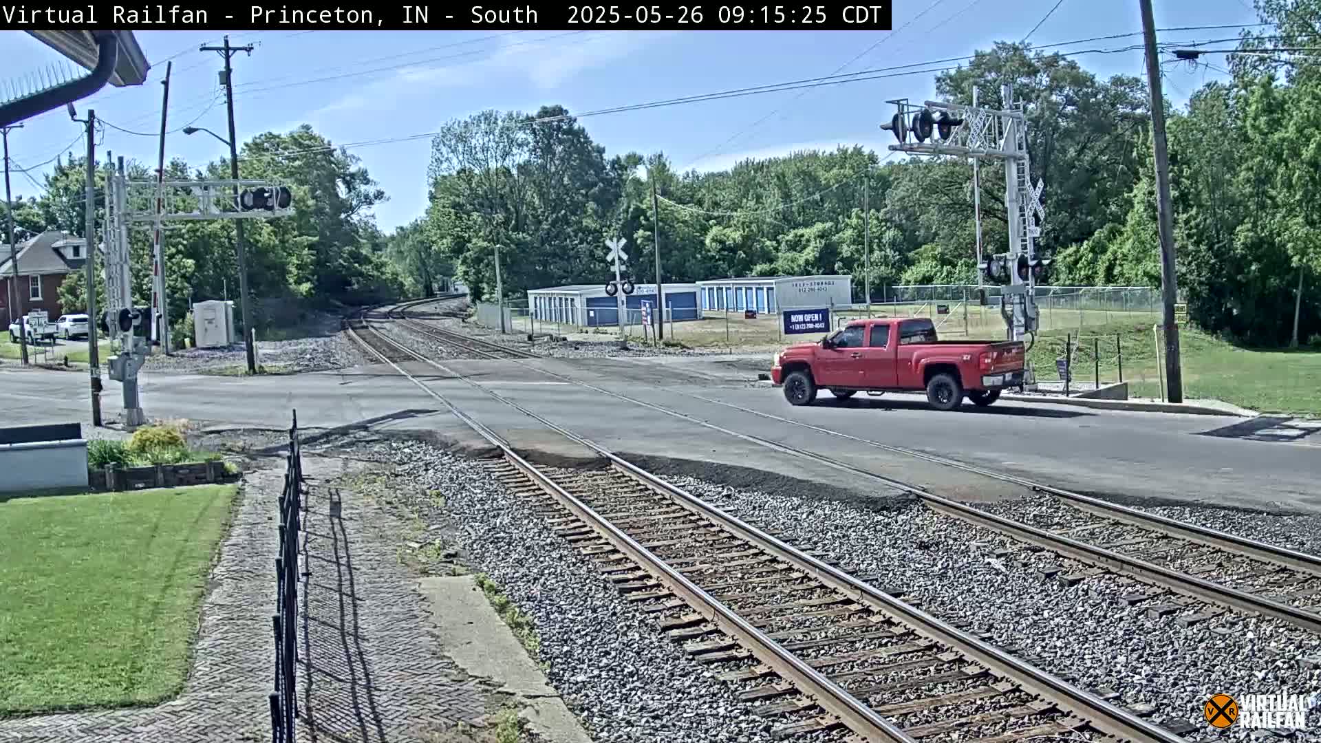 A red pickup truck is crossing a railroad intersection on a sunny day, with trees and buildings visible in the background.