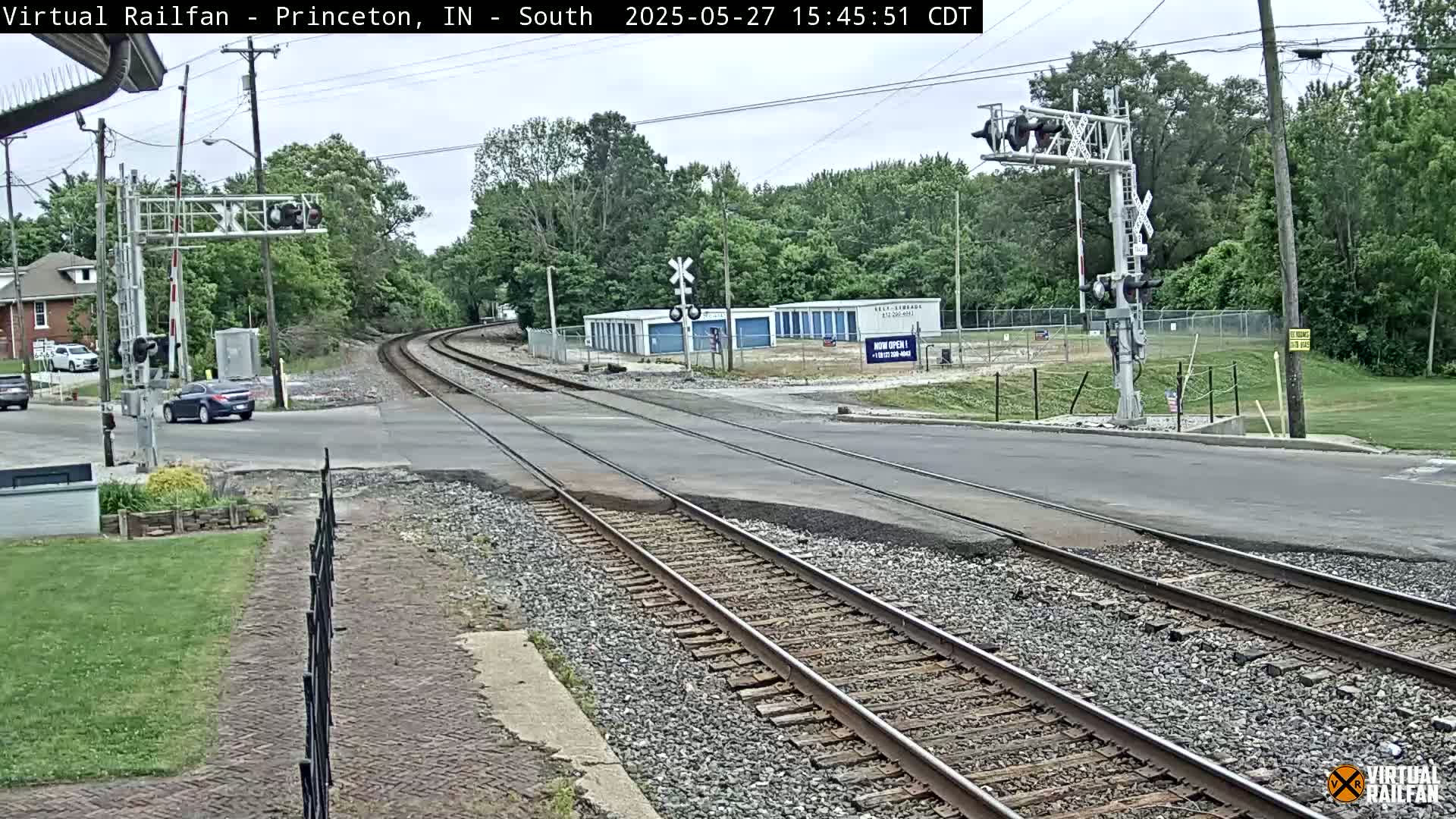 A railroad crossing with train tracks intersecting a road, under a partly cloudy sky, with trees and buildings in the background.