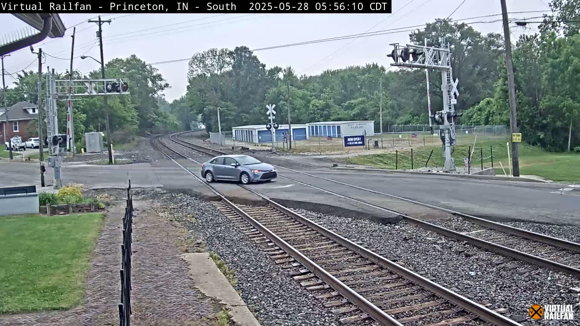 A silver car drives across a railroad crossing on a gray, overcast day, passing by storage units and a residential building.