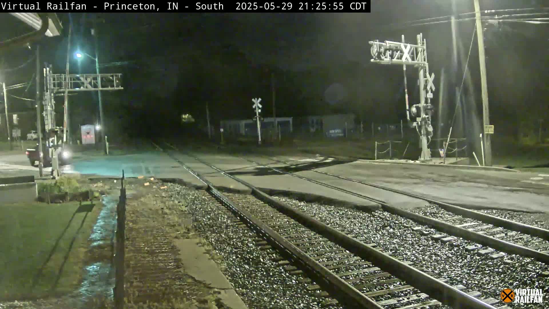A nighttime view of a railroad crossing with a roadway and train tracks, showing some damage to the road surface near the tracks.