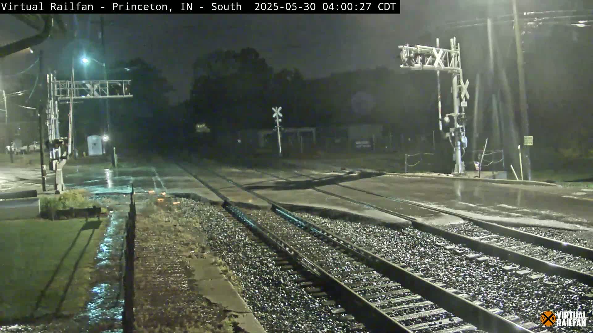 A nighttime view of rain-slicked railroad tracks and a road crossing under nighttime conditions.