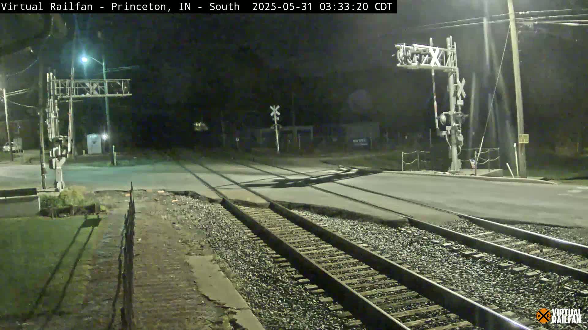 At night, a railroad crossing intersects a road under artificial light, with train tracks extending into the distance.
