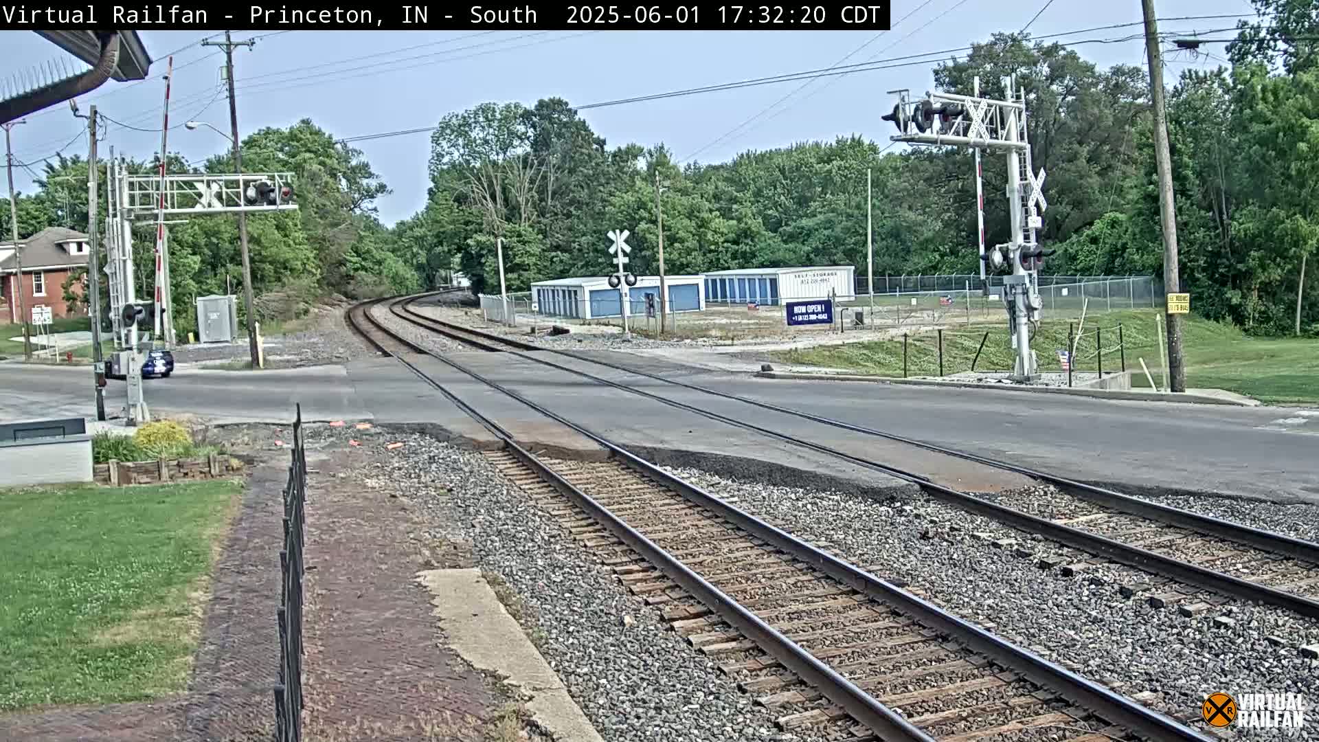 A mostly sunny view of a railroad crossing, with train tracks intersecting a road, surrounded by trees and buildings.