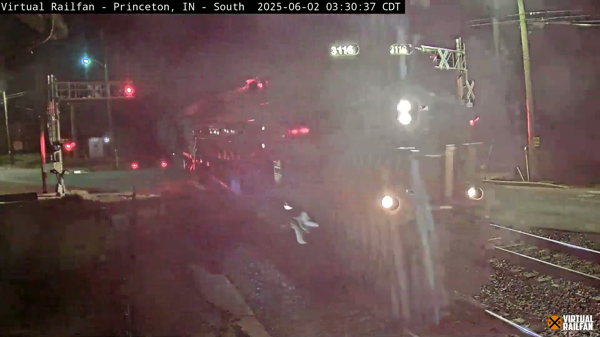 A dark-colored train passes a nighttime railroad crossing under rainy conditions.