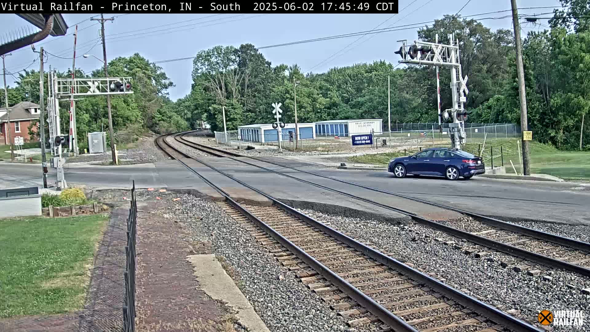 A dark-blue sedan drives across a railroad crossing under a mostly clear sky, past a row of storage units and railroad tracks that curve to the left.
