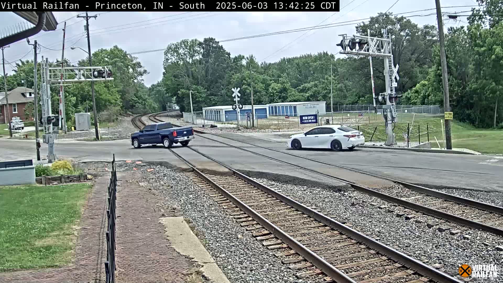 A blue pickup truck and a white sedan are crossing a railroad intersection under a partly cloudy sky.