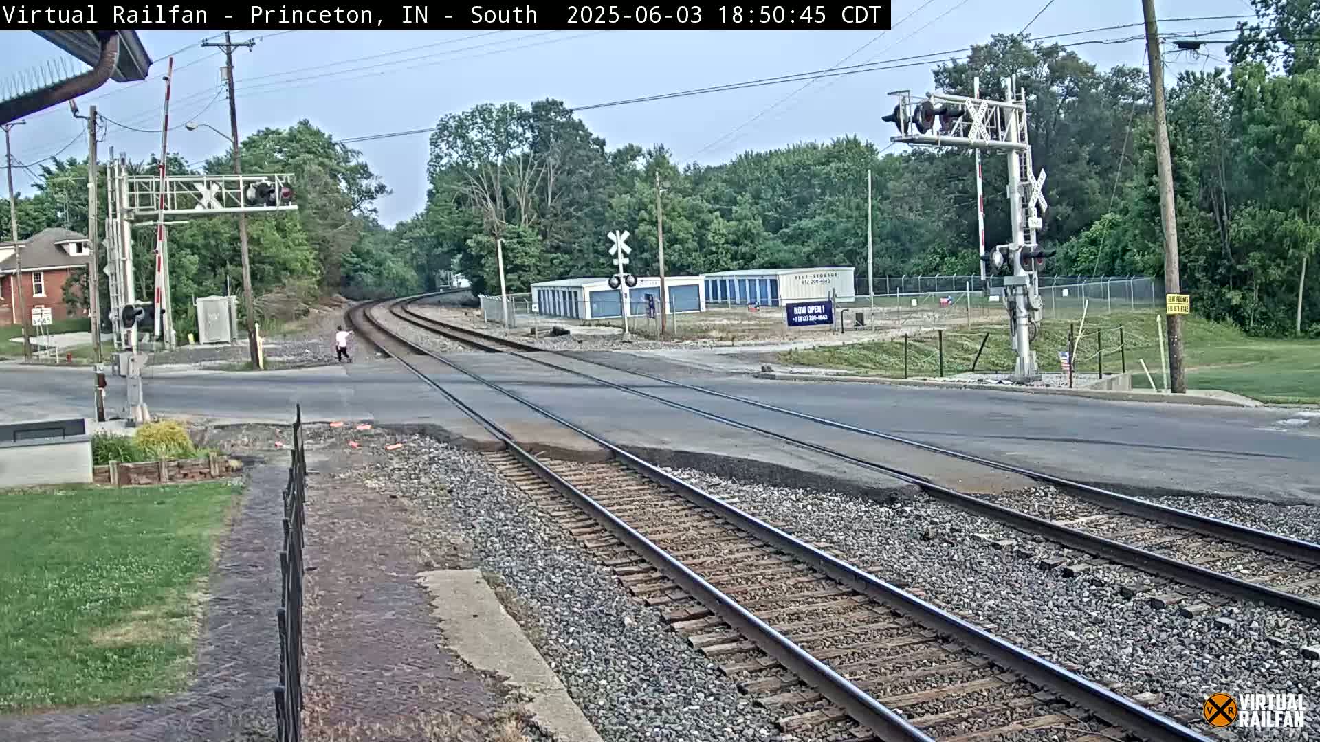 A partly cloudy daytime view of a railroad crossing with a person walking across the tracks near a street and several storage units.