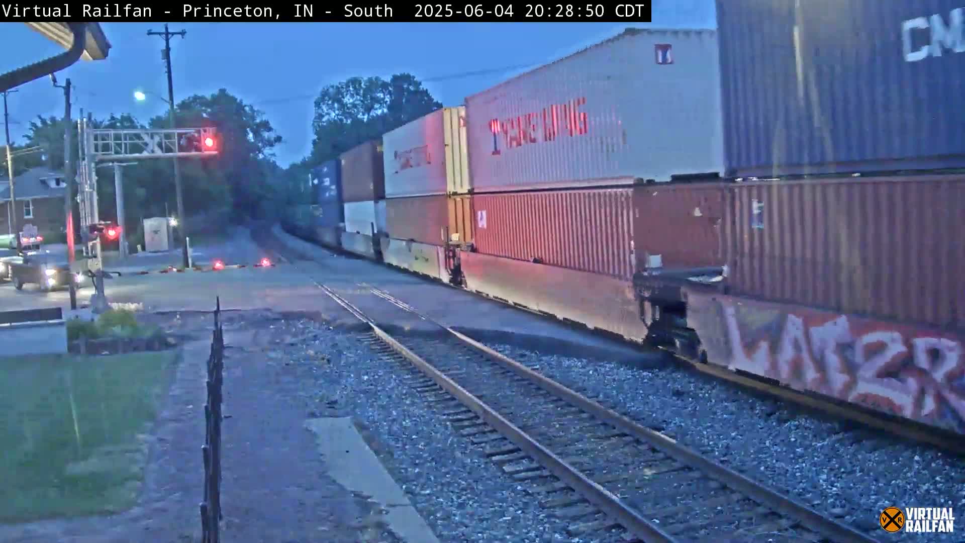A long freight train passes a road crossing at dusk under a mostly clear sky.