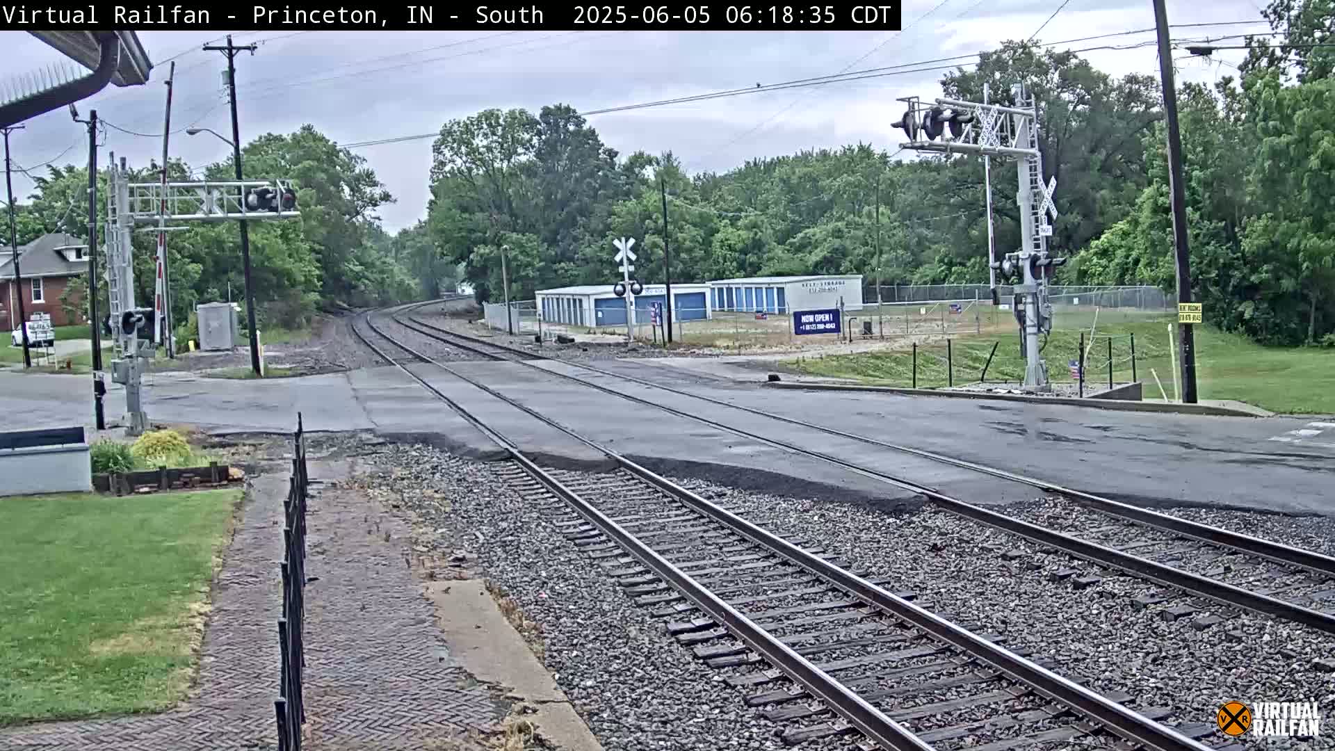 A railroad crossing with train tracks intersecting a road, under an overcast sky, with trees and buildings in the background.