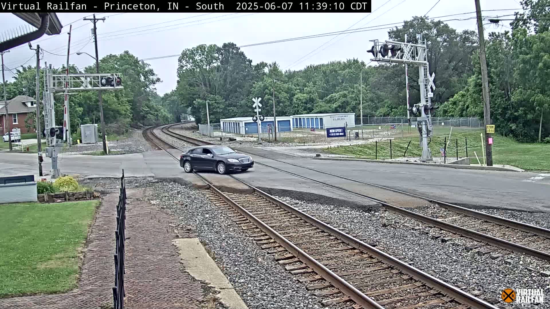 A dark-colored sedan crosses a railroad track at a road intersection on an overcast day.