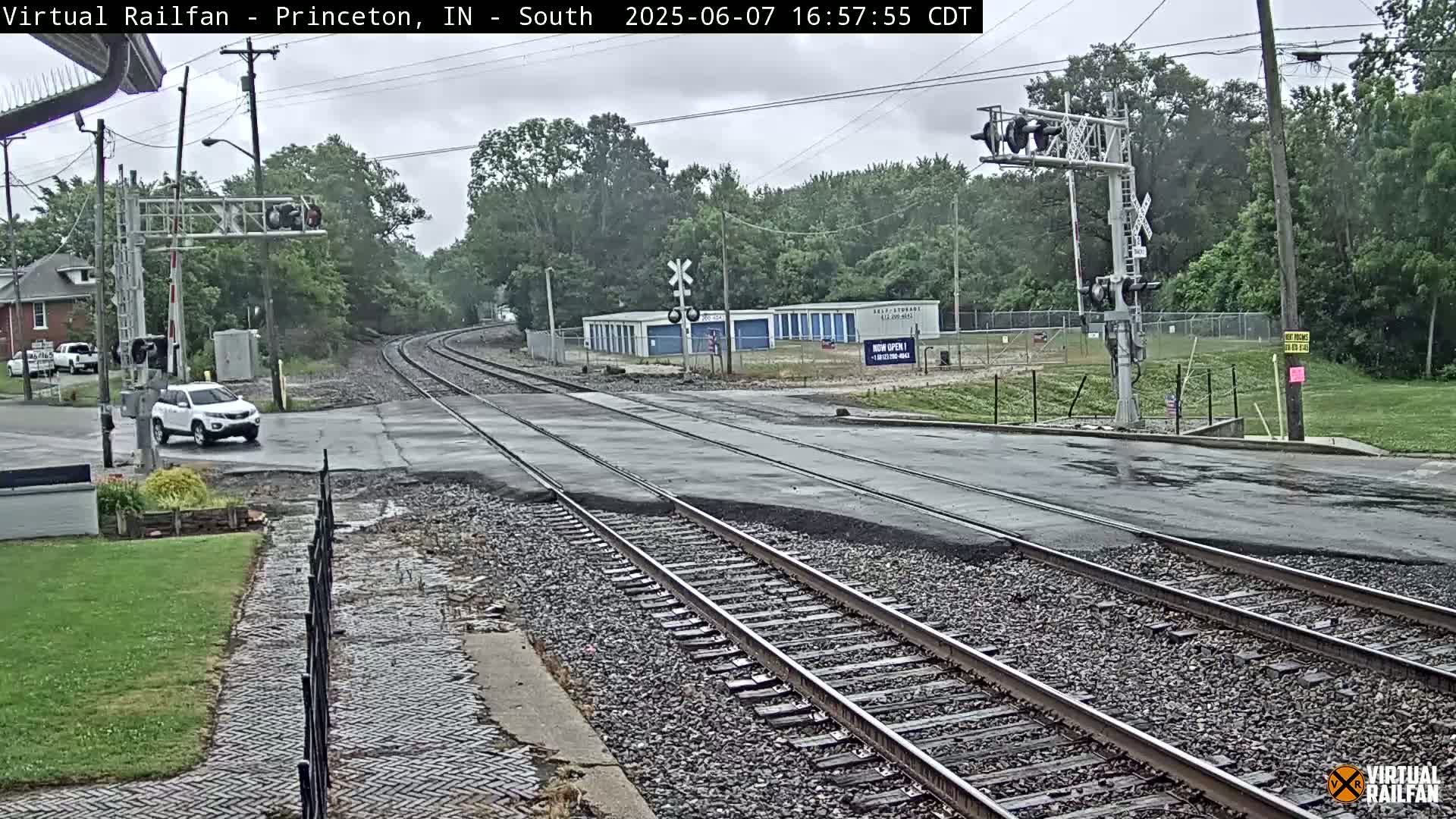 A white SUV drives across a railroad crossing on a wet, overcast day, with railroad tracks extending into a tree-lined background.