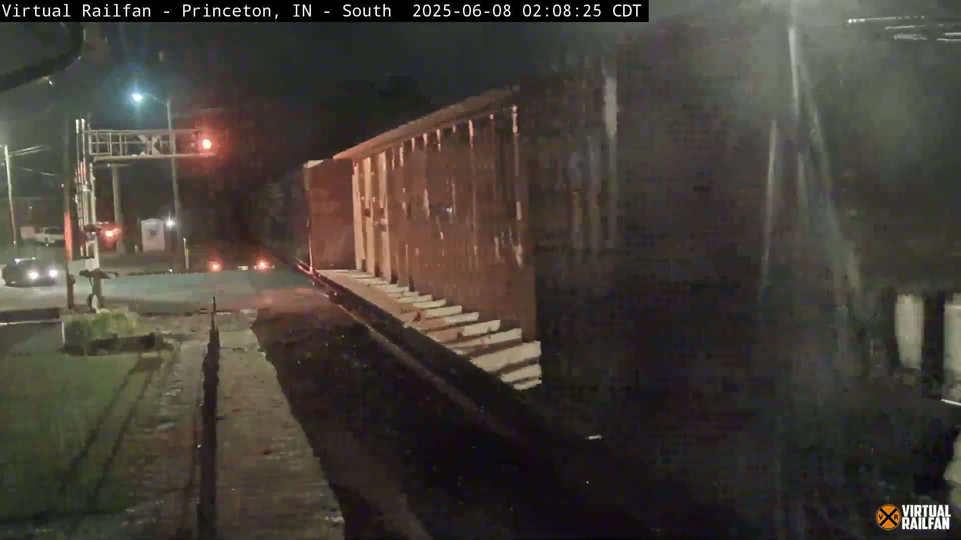 At night, a long freight train passes a road crossing under a red traffic light, with a person walking nearby.