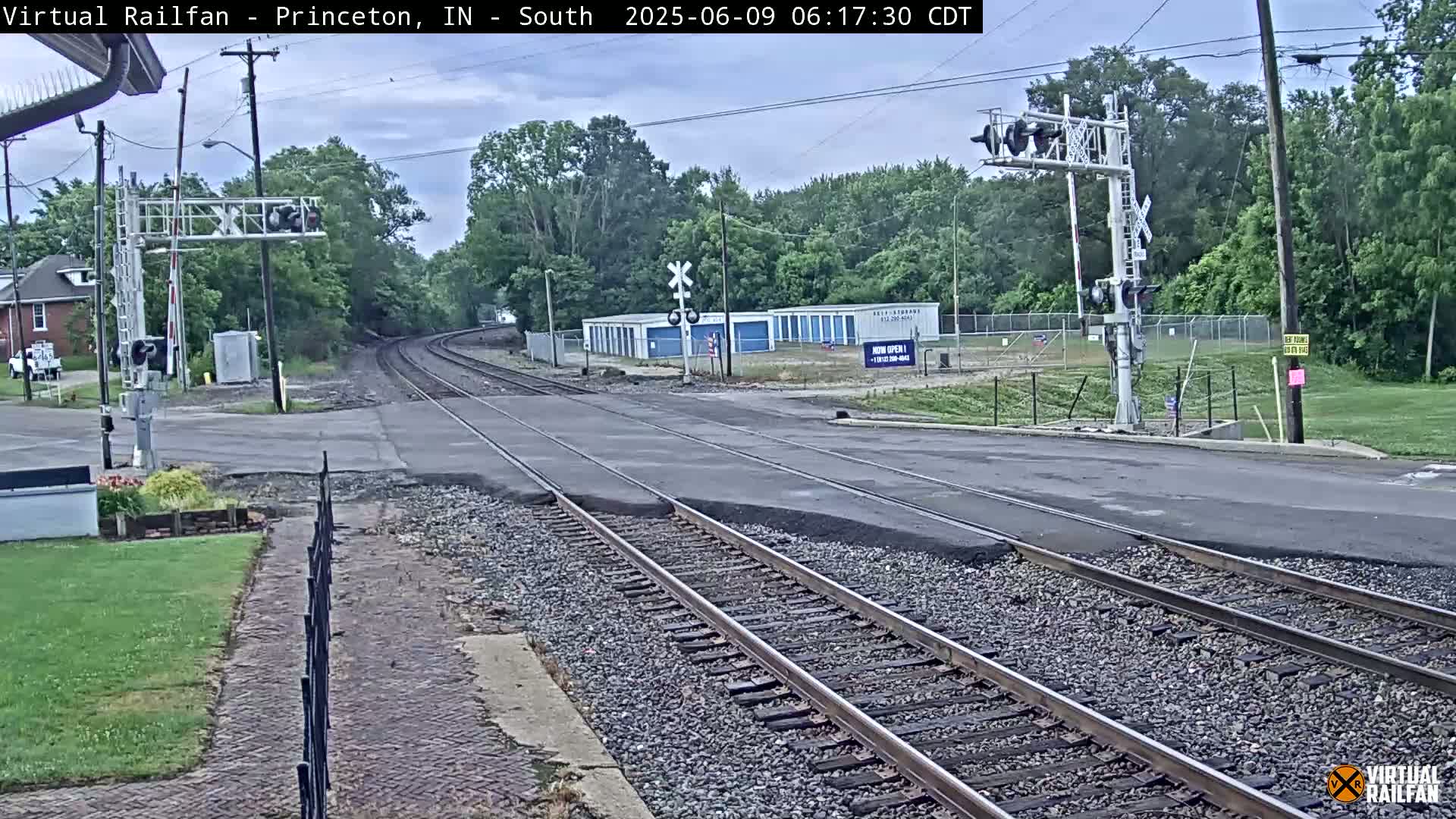 A railroad crossing with train tracks intersecting a road, under an overcast sky, is surrounded by trees and a few buildings.