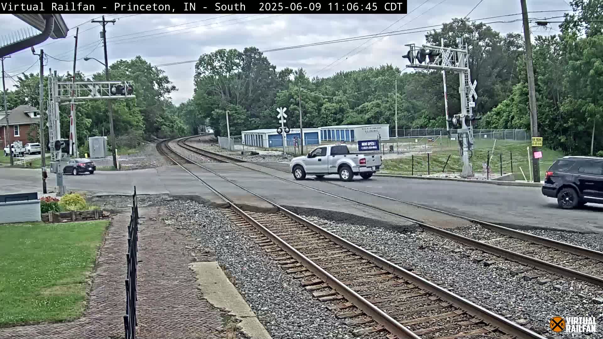 A lightly overcast day shows a road crossing railroad tracks, with a pickup truck and SUV driving across the intersection near railroad crossing signals and some buildings in the background.
