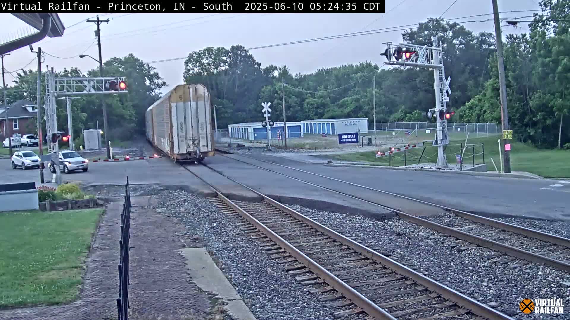 A long freight train passes a road crossing under a twilight sky, with several cars waiting at the crossing.