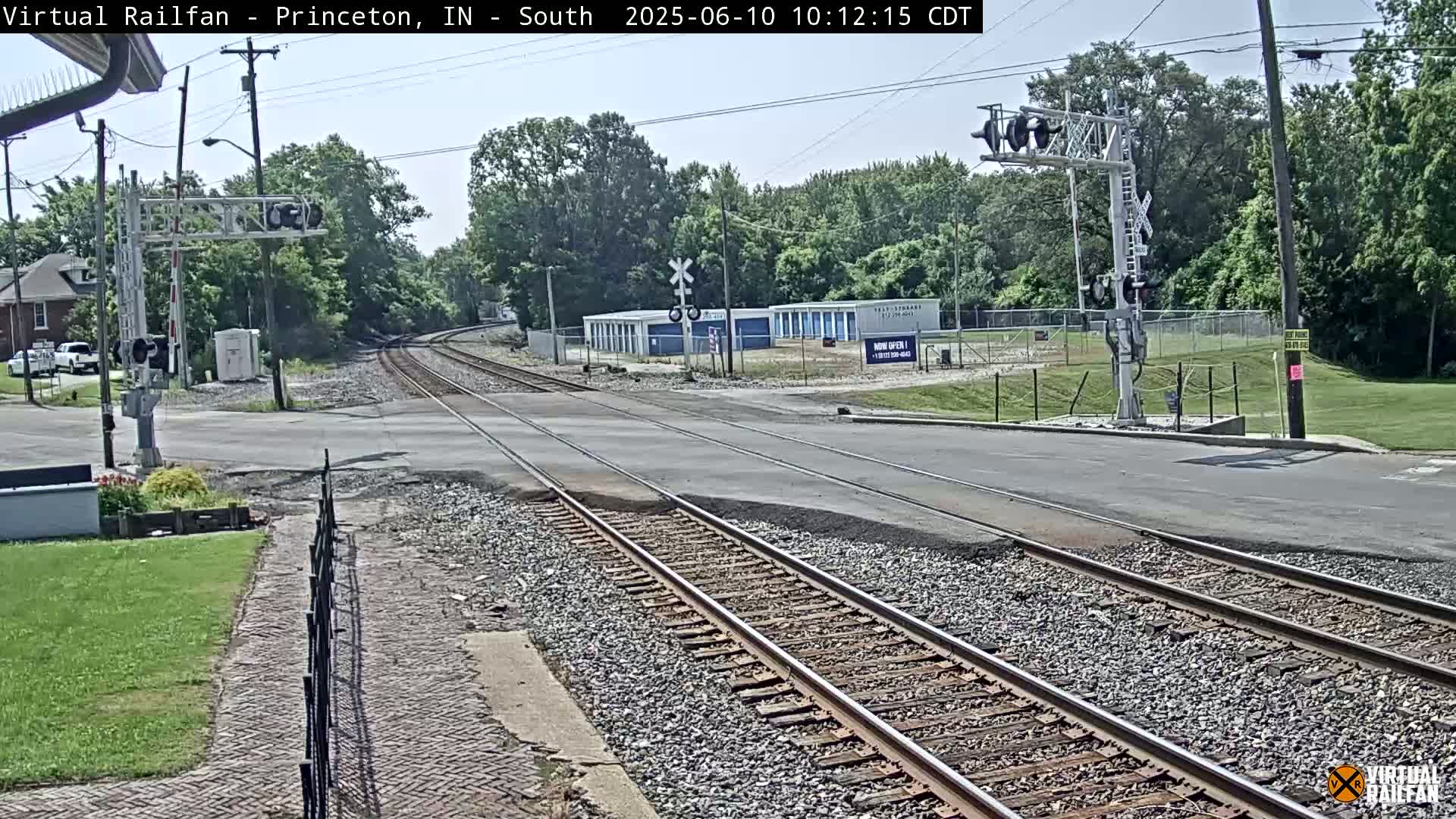 A sunny day shows railroad tracks intersecting a road, with a signal tower and storage units visible in the background.