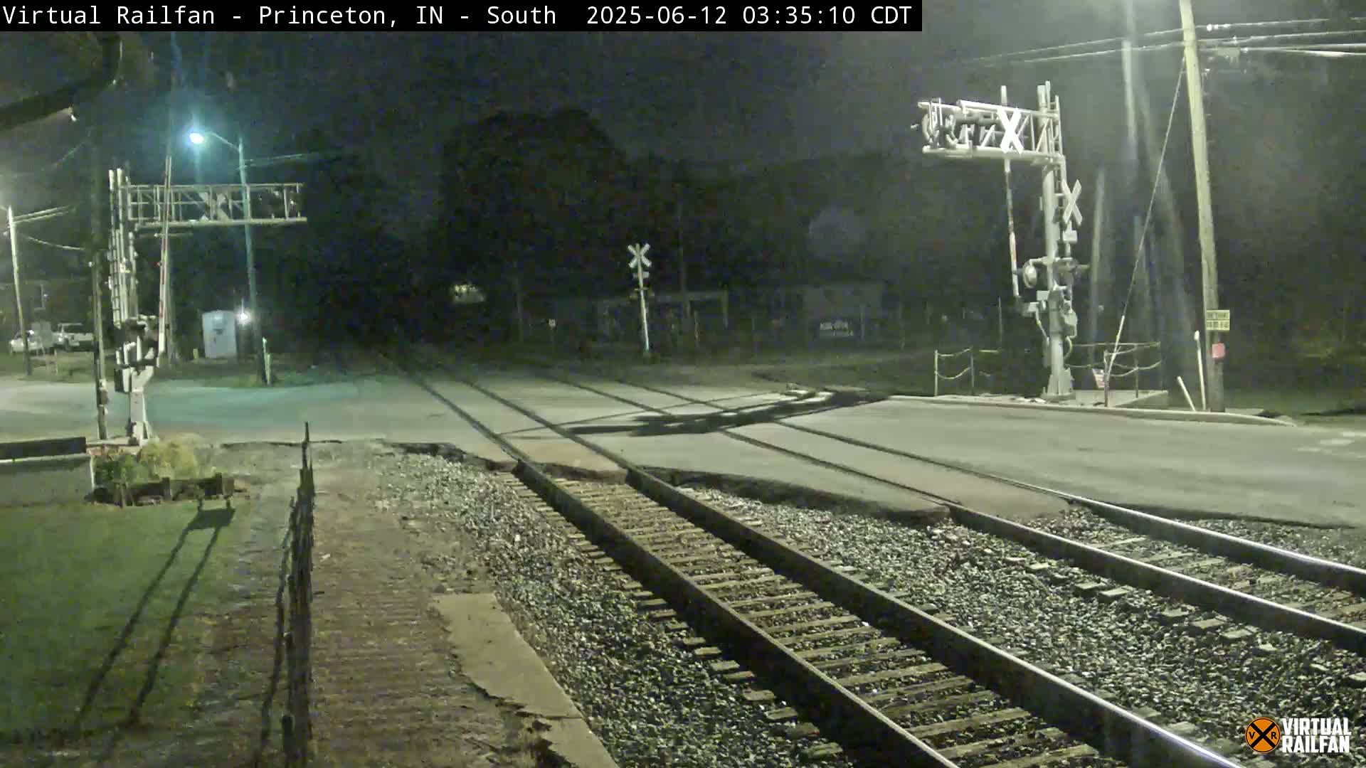 A nighttime view of a railroad crossing with train tracks intersecting a road under artificial light.