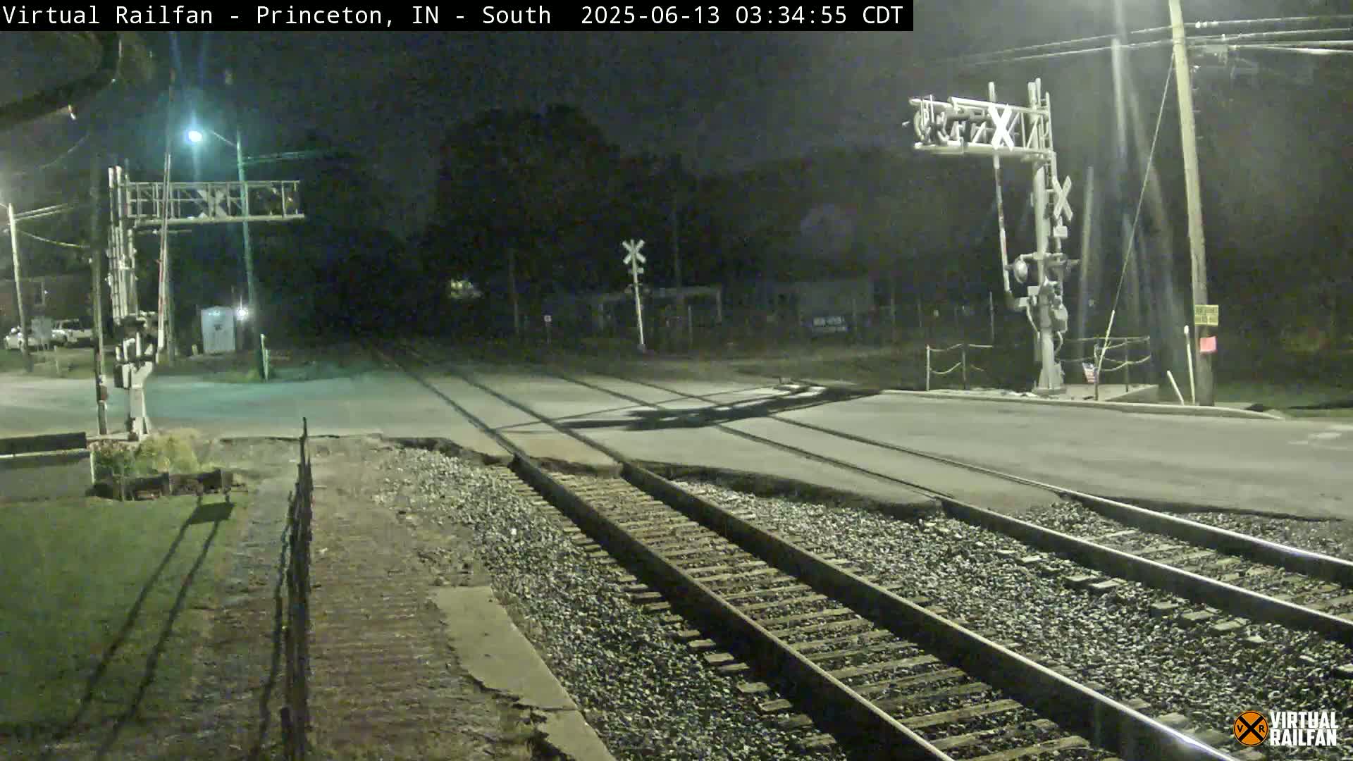 A nighttime view of a railroad crossing with train tracks intersecting a road under artificial light.