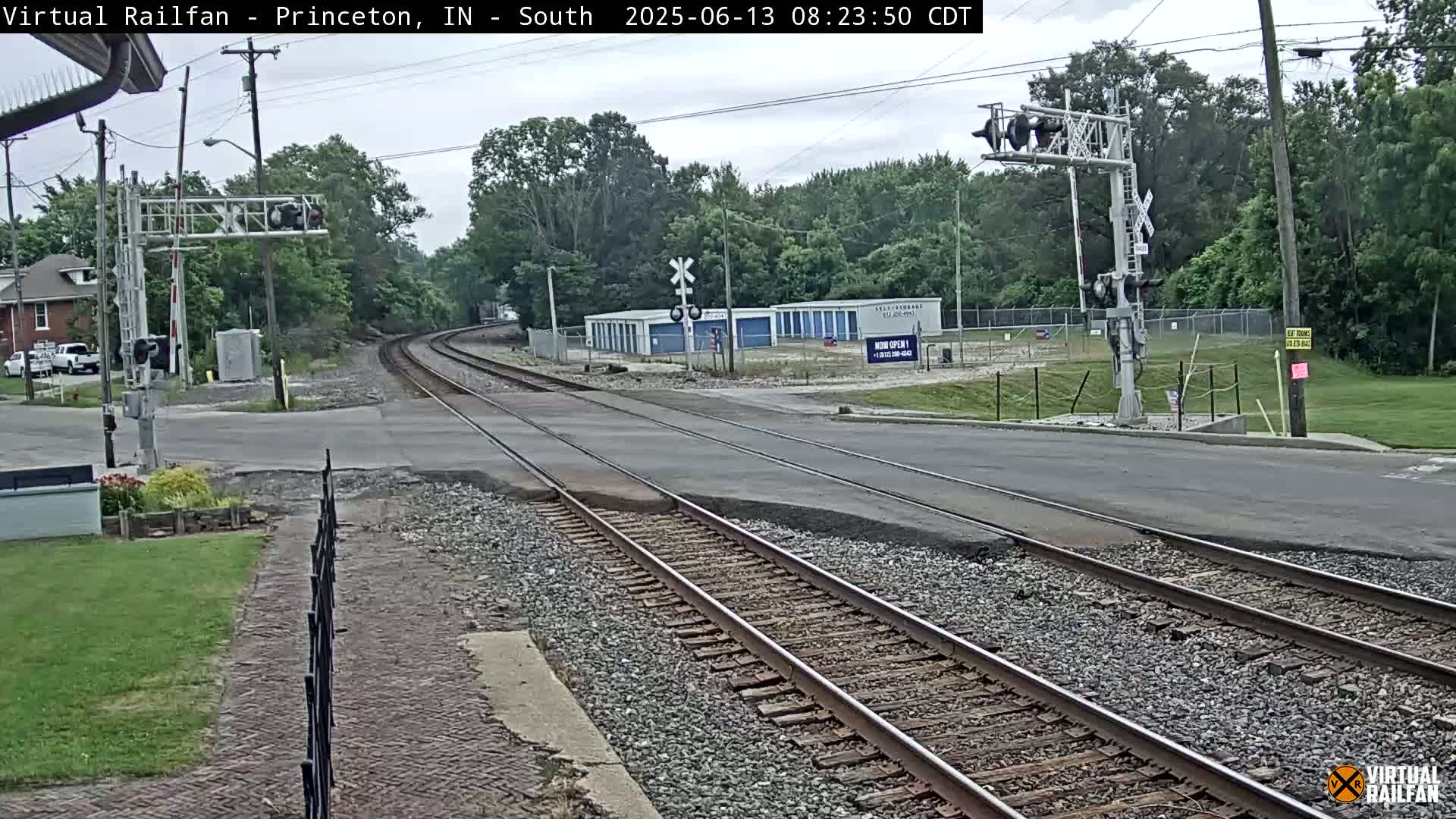 Railroad tracks cross a road under an overcast sky, passing in front of a row of storage units and some trees.