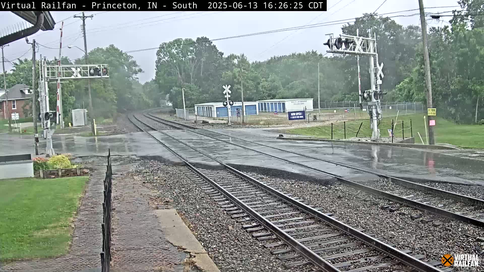A wet railroad crossing in light rain shows train tracks intersecting a road under a mostly cloudy sky, with trees and buildings visible in the background.
