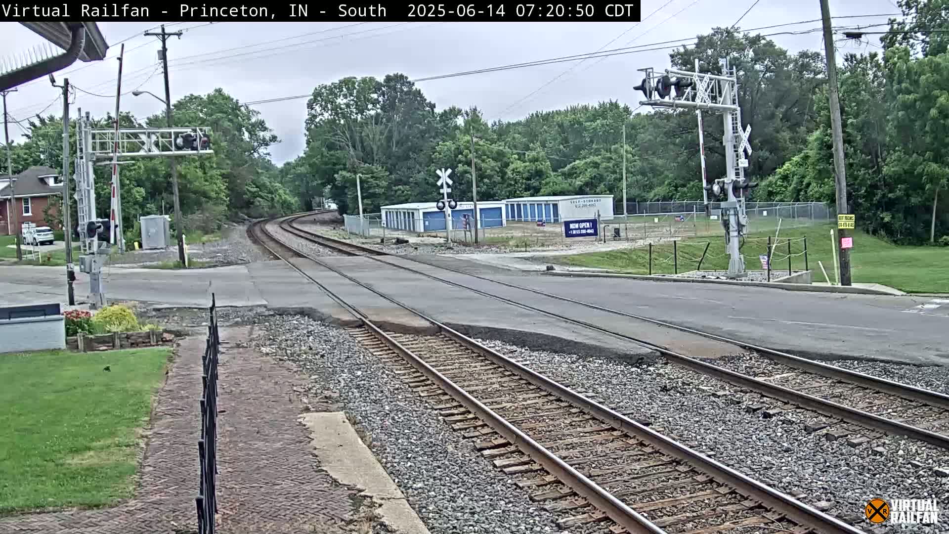 A railroad crossing with train tracks intersecting a road under an overcast sky, surrounded by trees and some buildings in the background.