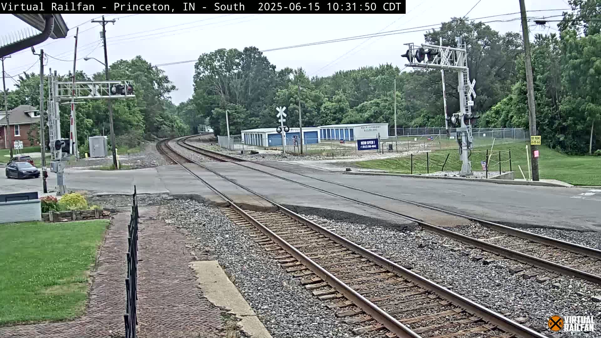 A railroad crossing with train tracks intersecting a road under an overcast sky, surrounded by trees and buildings.