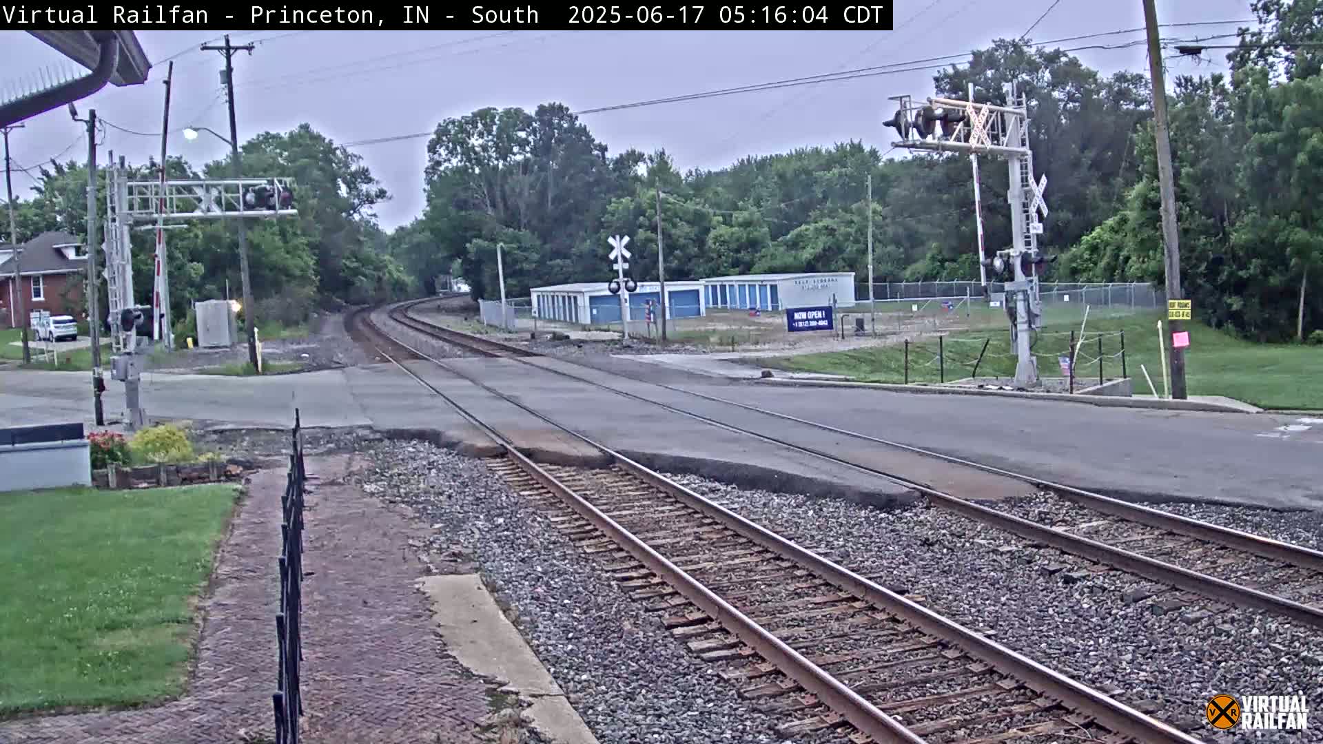 A railroad crossing with train tracks intersecting a road, under an overcast sky, with trees and buildings visible in the background.