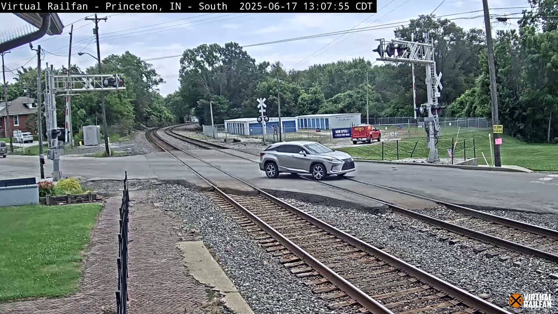 A silver SUV drives across a railroad crossing in a mostly sunny area with green trees and buildings in the background.