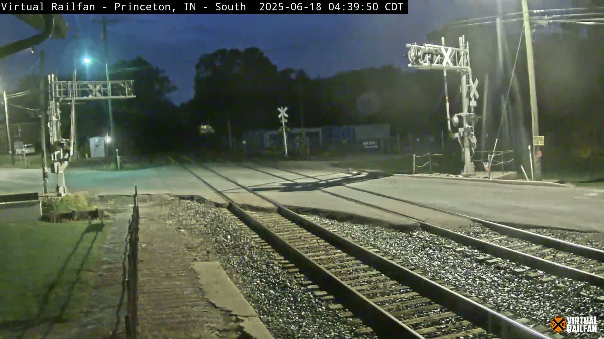 A nighttime view of a railroad crossing with train tracks and a road intersecting, under a dark sky.