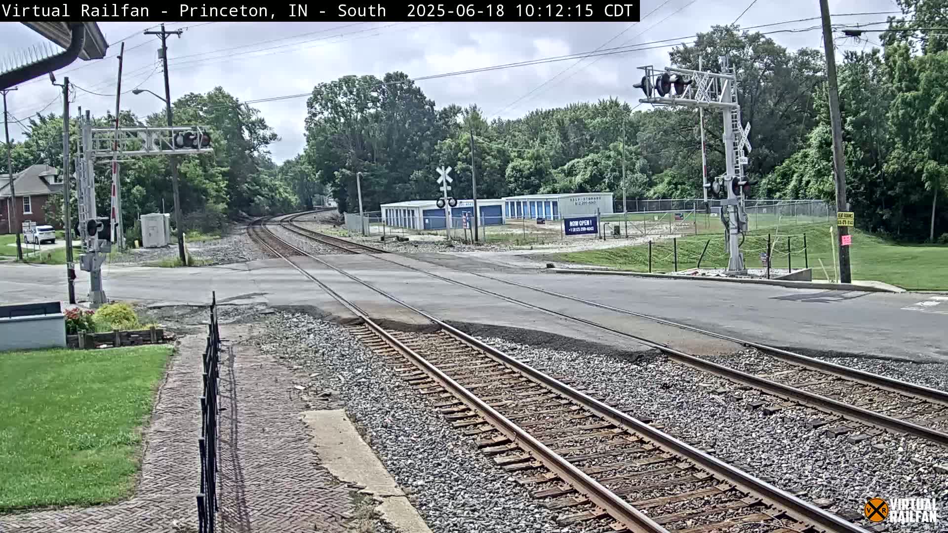 A railroad crossing with train tracks intersecting a road under a partly cloudy sky, showing a row of storage units and trees in the background.