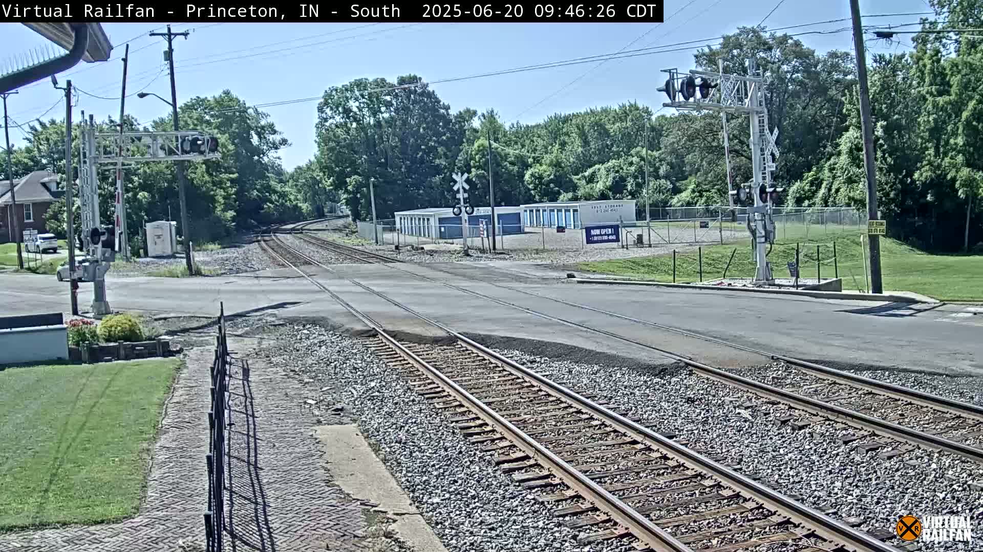 A sunny day shows a railroad crossing with train tracks intersecting a road, surrounded by trees and buildings.