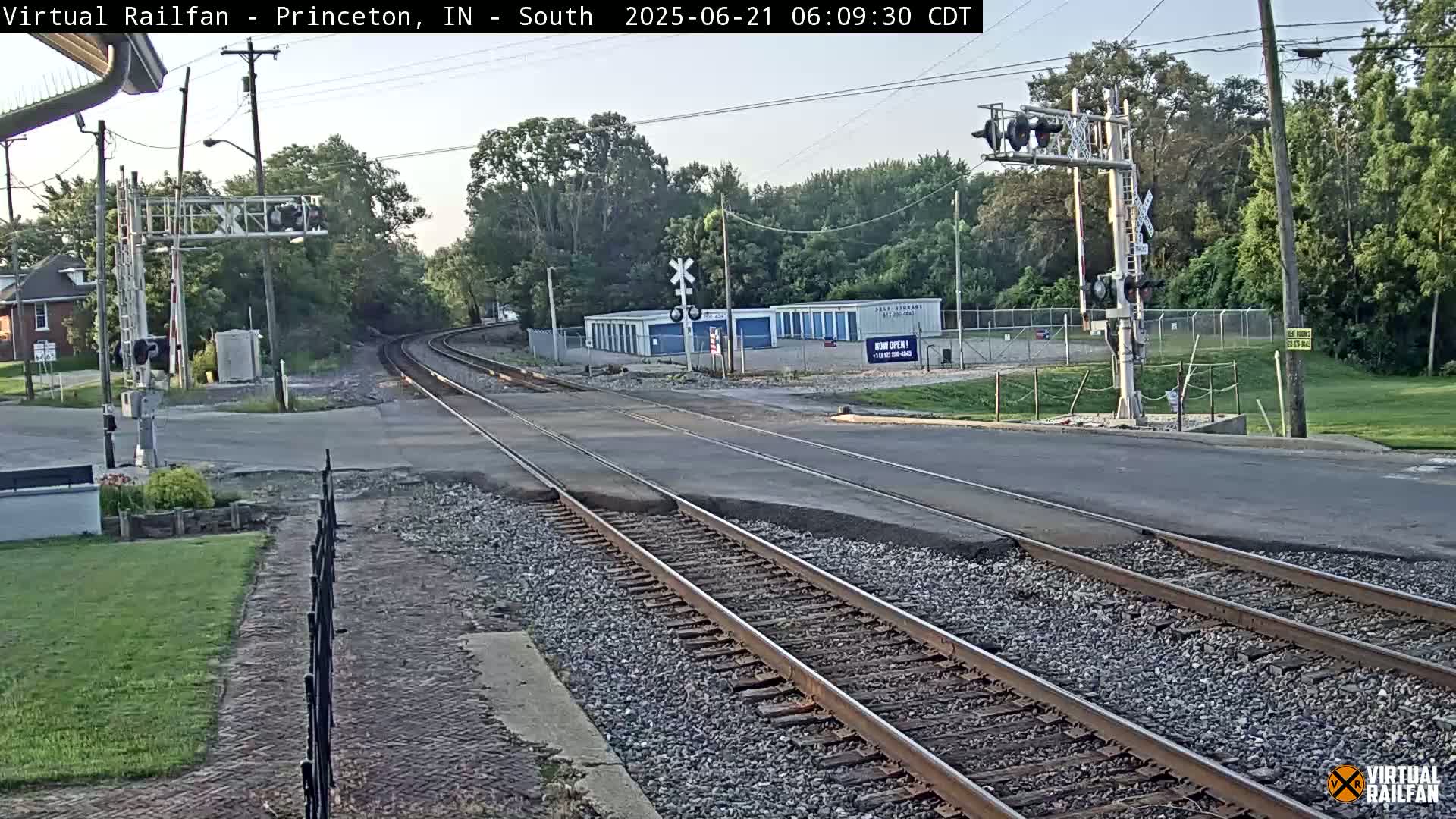 Train tracks intersect a road near a row of storage units under a partly cloudy sky.