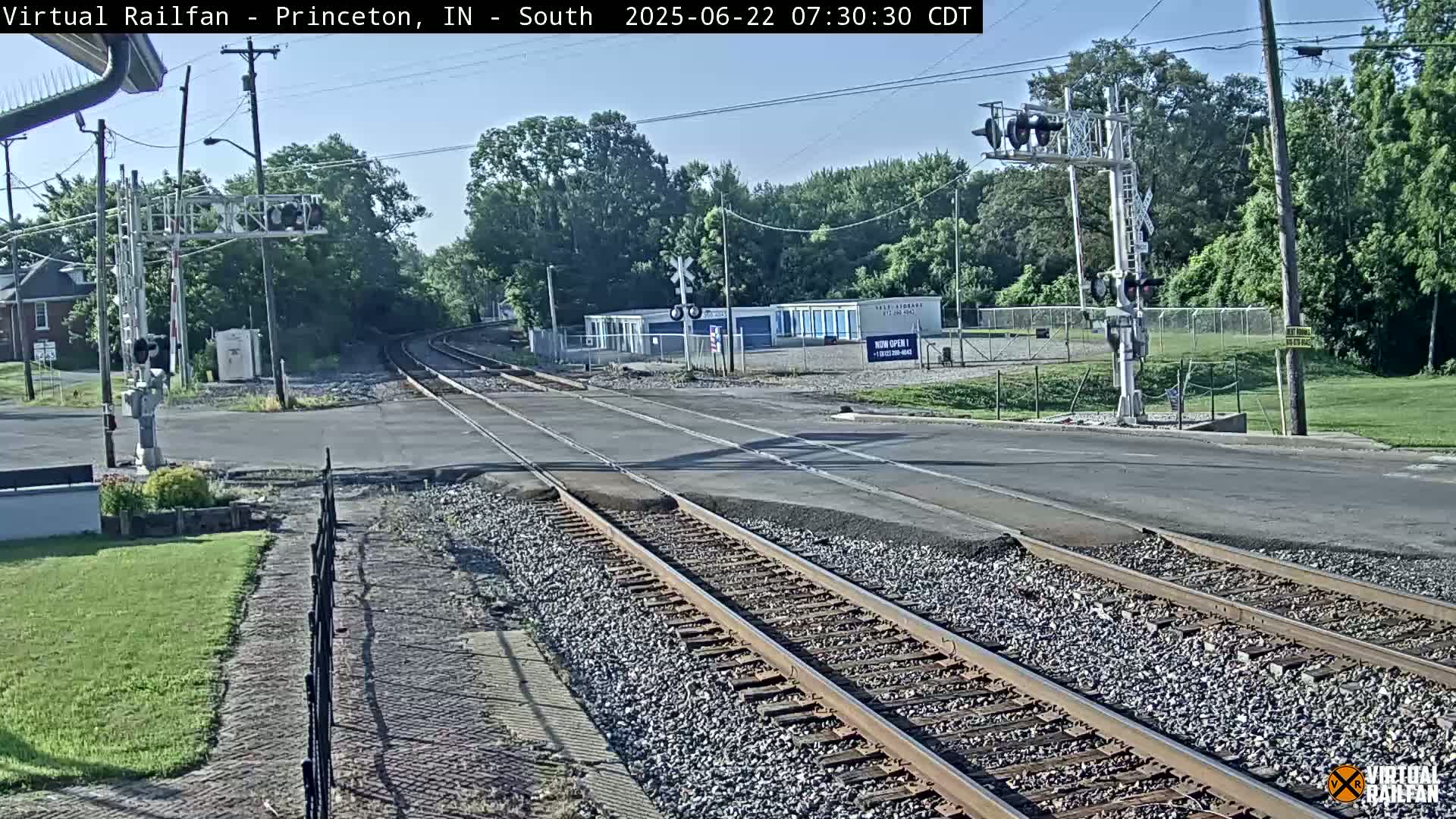 A railroad crossing with train tracks intersecting a road, under a mostly clear sky, surrounded by green trees and grass.