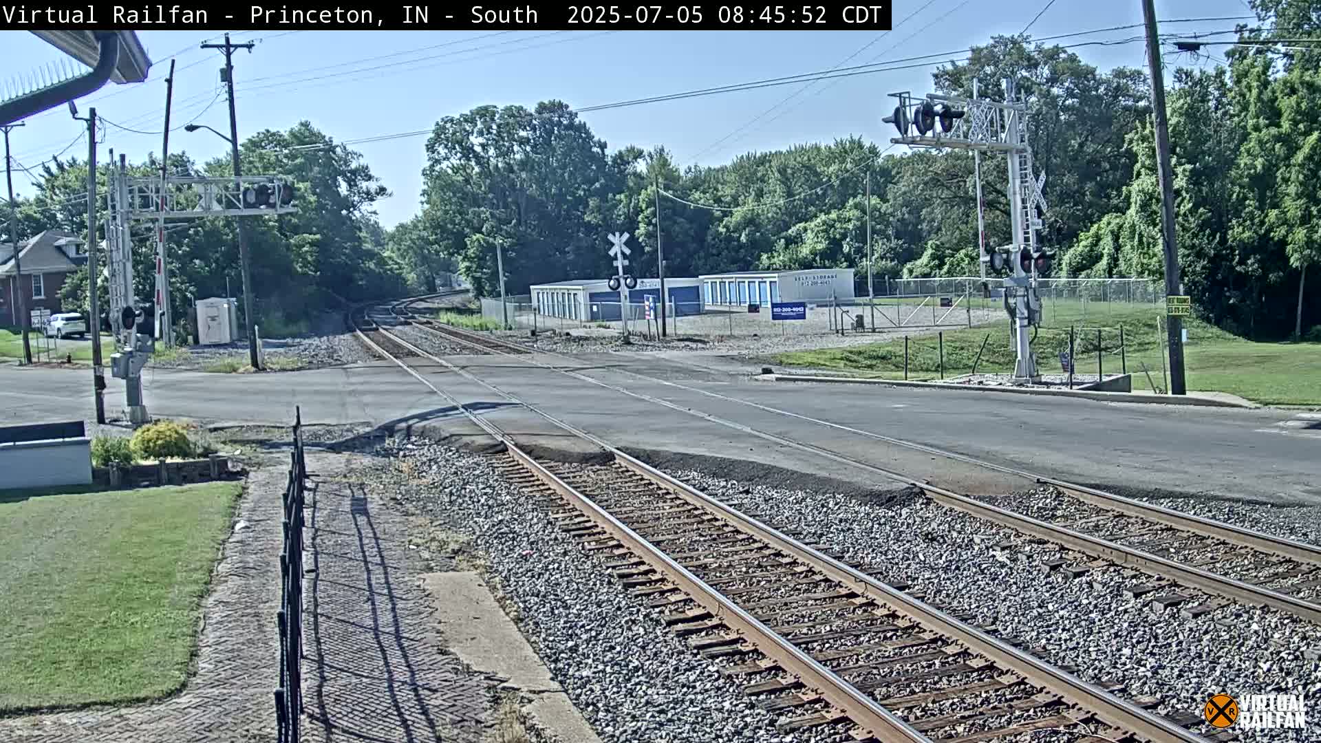 A sunny day shows railroad tracks crossing a road, with a row of storage units visible in the background.