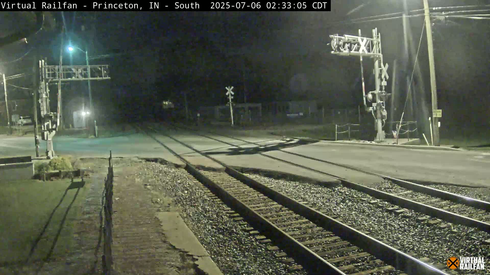A nighttime view of a railroad crossing with two sets of train tracks intersecting a paved road, under dark conditions.