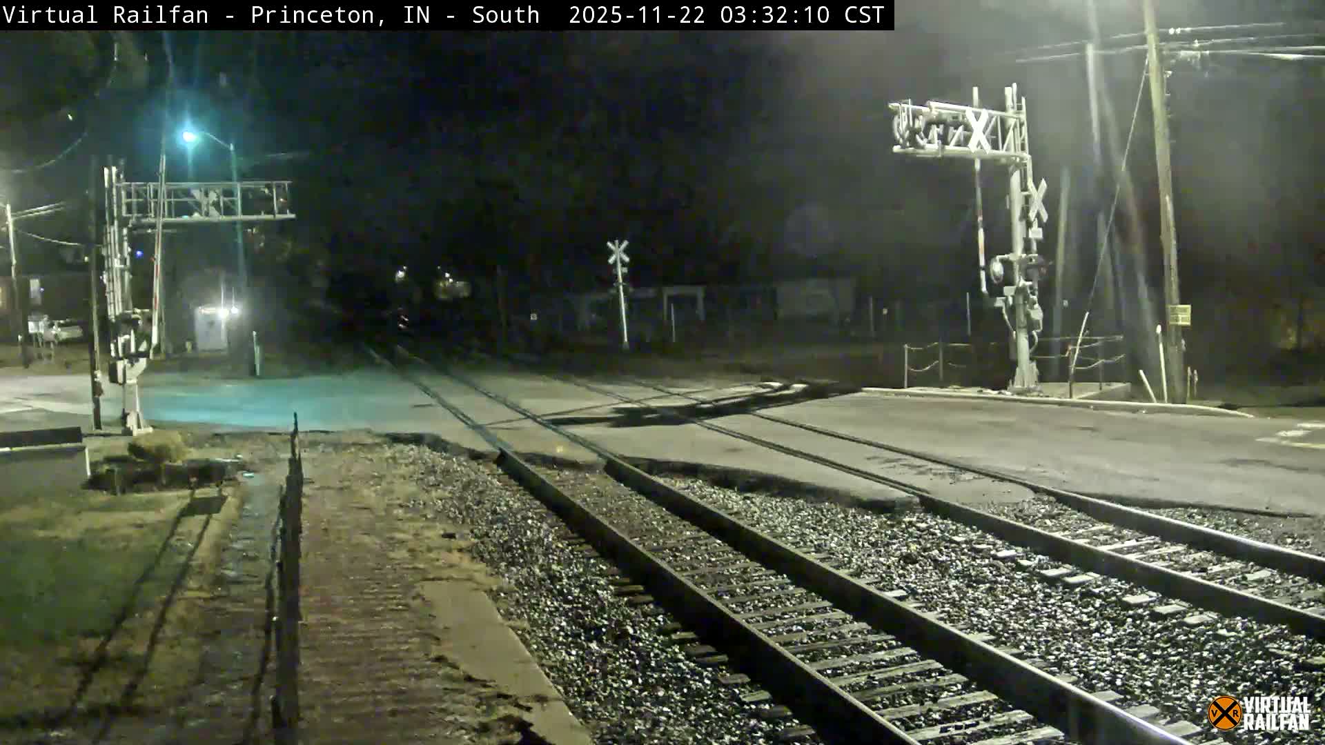 A clear night captures a multi-track railroad crossing a paved road, marked by signals and illuminated by streetlights.