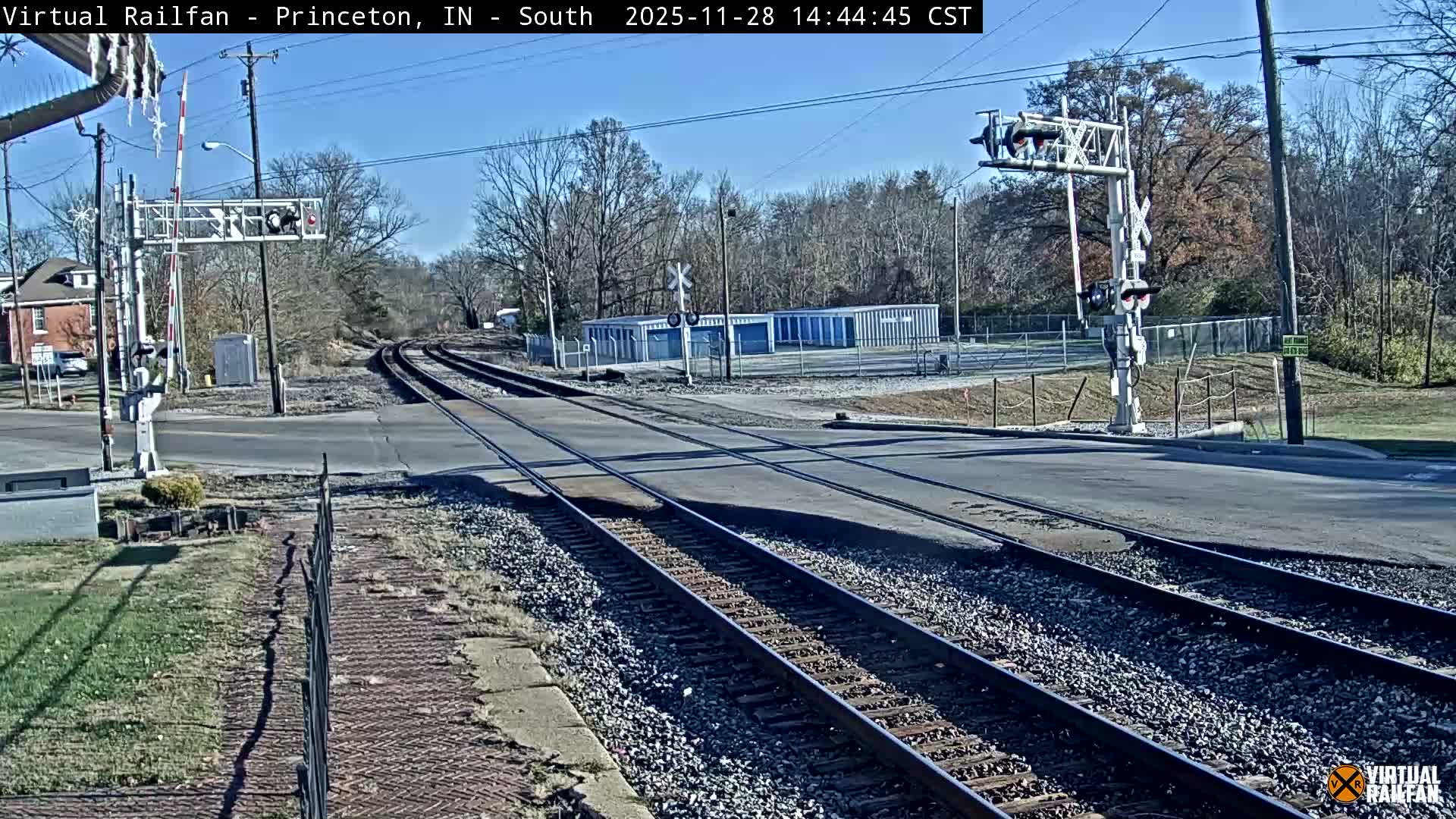A railroad crossing features multiple tracks intersecting an asphalt road, flanked by bare trees, residential buildings, and storage units, all under a clear, sunny blue sky.