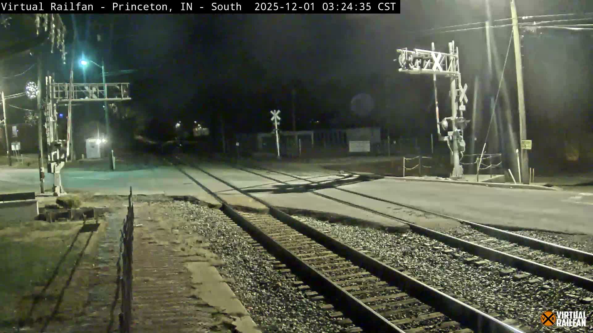 A clear night view captures multiple railroad tracks converging at a paved road crossing, illuminated by streetlights and featuring prominent railroad crossing signals.