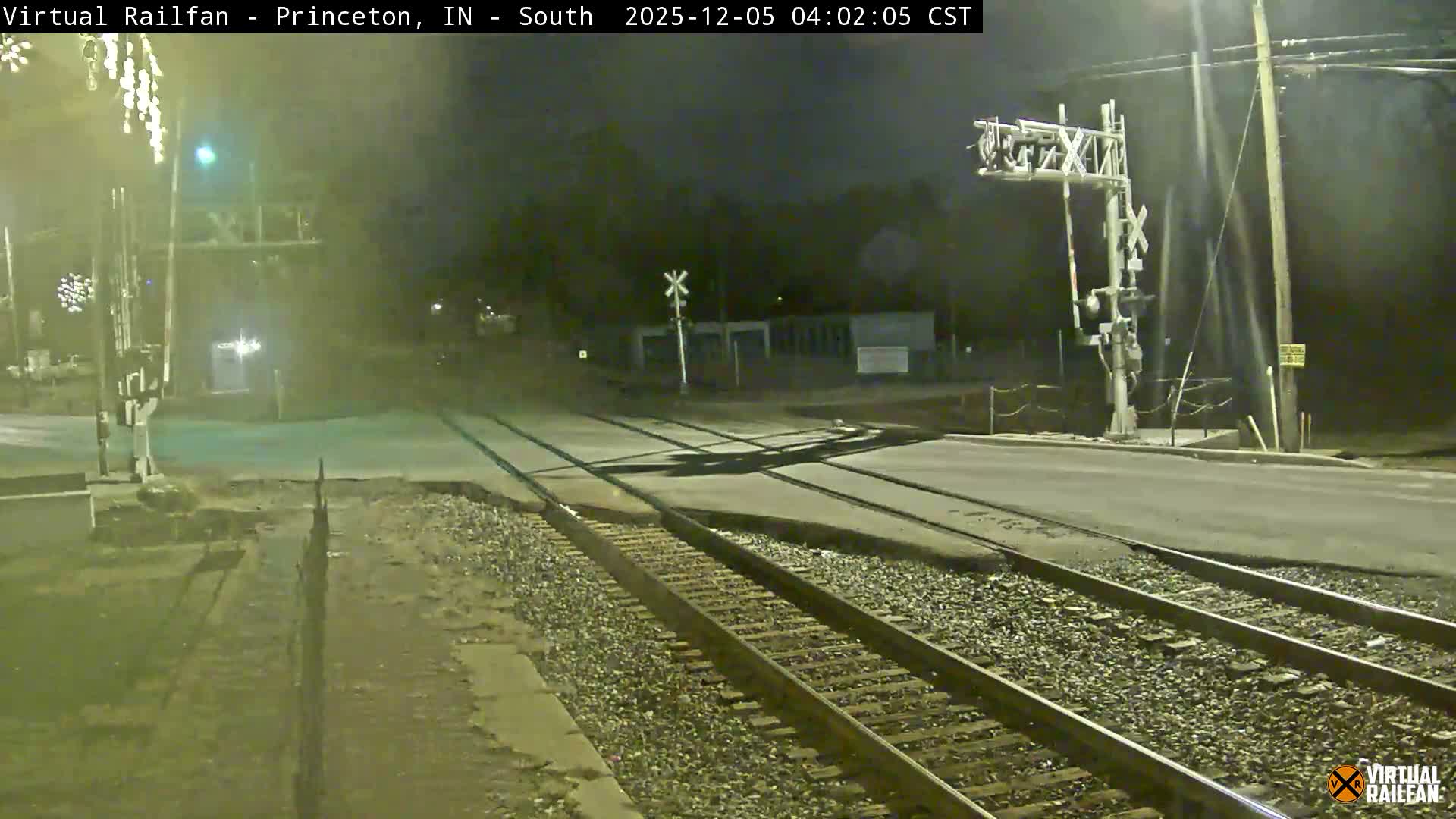 A nighttime view reveals a railroad crossing with multiple tracks intersecting a paved road, illuminated by various streetlights and crossing signals under clear skies.