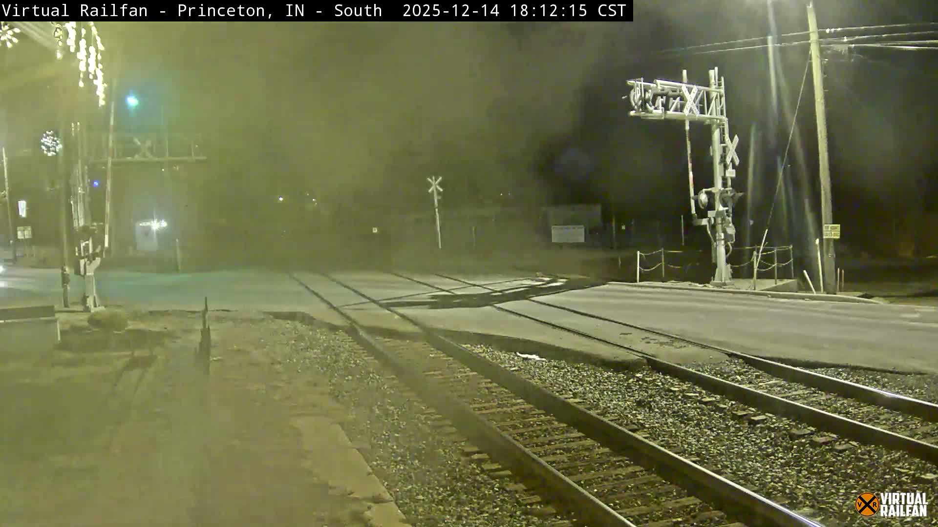 A nighttime view reveals a railroad crossing with multiple tracks intersecting a paved road, illuminated by various streetlights and crossing signals under clear skies.